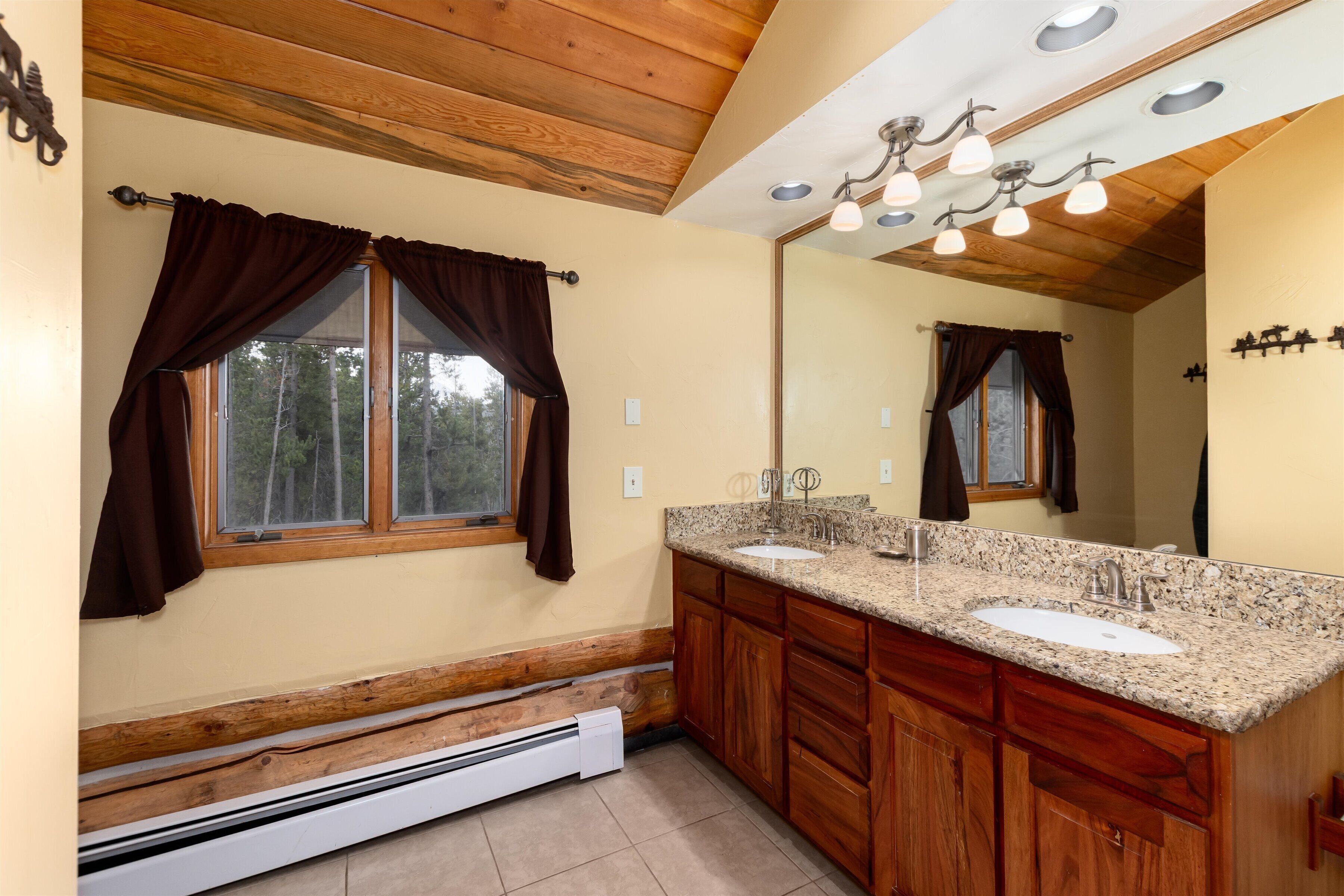 A bathroom with wooden cabinets, a granite countertop, a double sink, a large mirror, and a ceiling light fixture.
