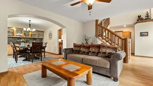 A cozy living room with a wooden coffee table, a gray sofa with patterned cushions, a wooden staircase, and a dining area with a chandelier. - Huge, Family Friendly, 6BR Retreat! Sleeps 16! All King Beds. Hot Tub and Sauna! (Winter Park)