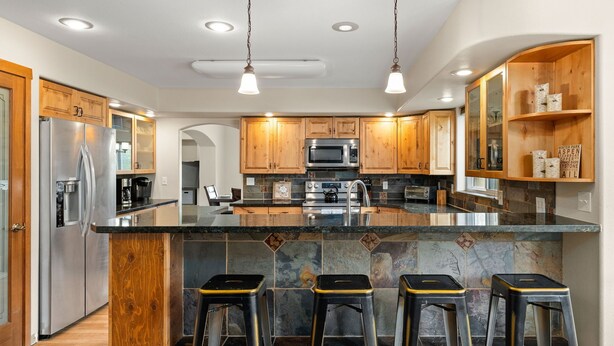 A modern kitchen with wooden cabinets, stainless steel appliances, a large island with stools, and a stone backsplash. - Huge, Family Friendly, 6BR Retreat! Sleeps 16! All King Beds. Hot Tub and Sauna! (Winter Park)