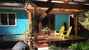 Une terrasse en bois avec un fauteuil Adirondack jaune, un patio couvert et un bâtiment bleu doté d’une fenêtre et d’une porte.