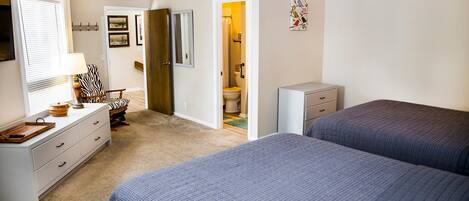 A bedroom with two beds, a dresser, a chair, and a bathroom visible through an open door.