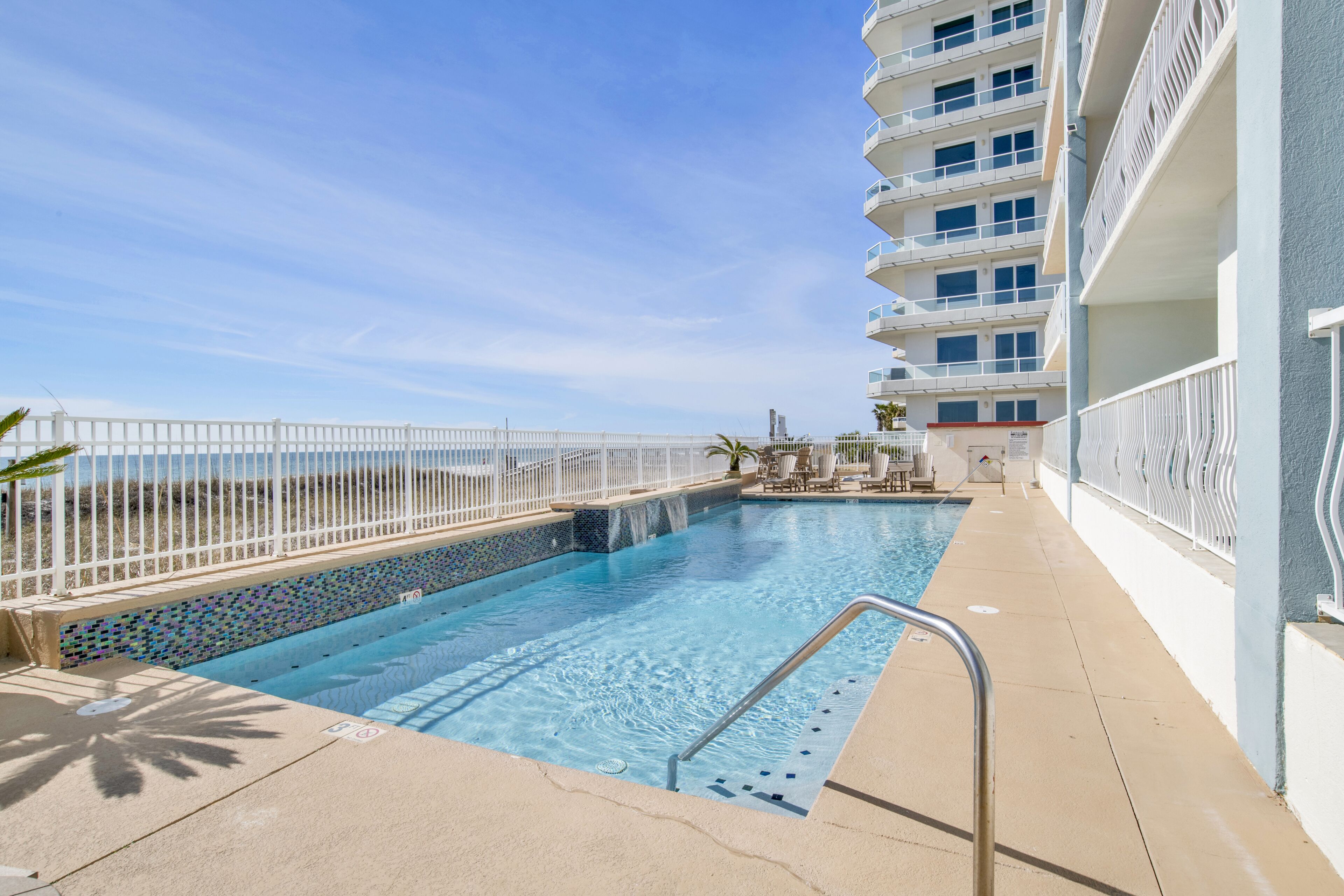 A swimming pool with a white fence, a clear blue sky, and a building with balconies.