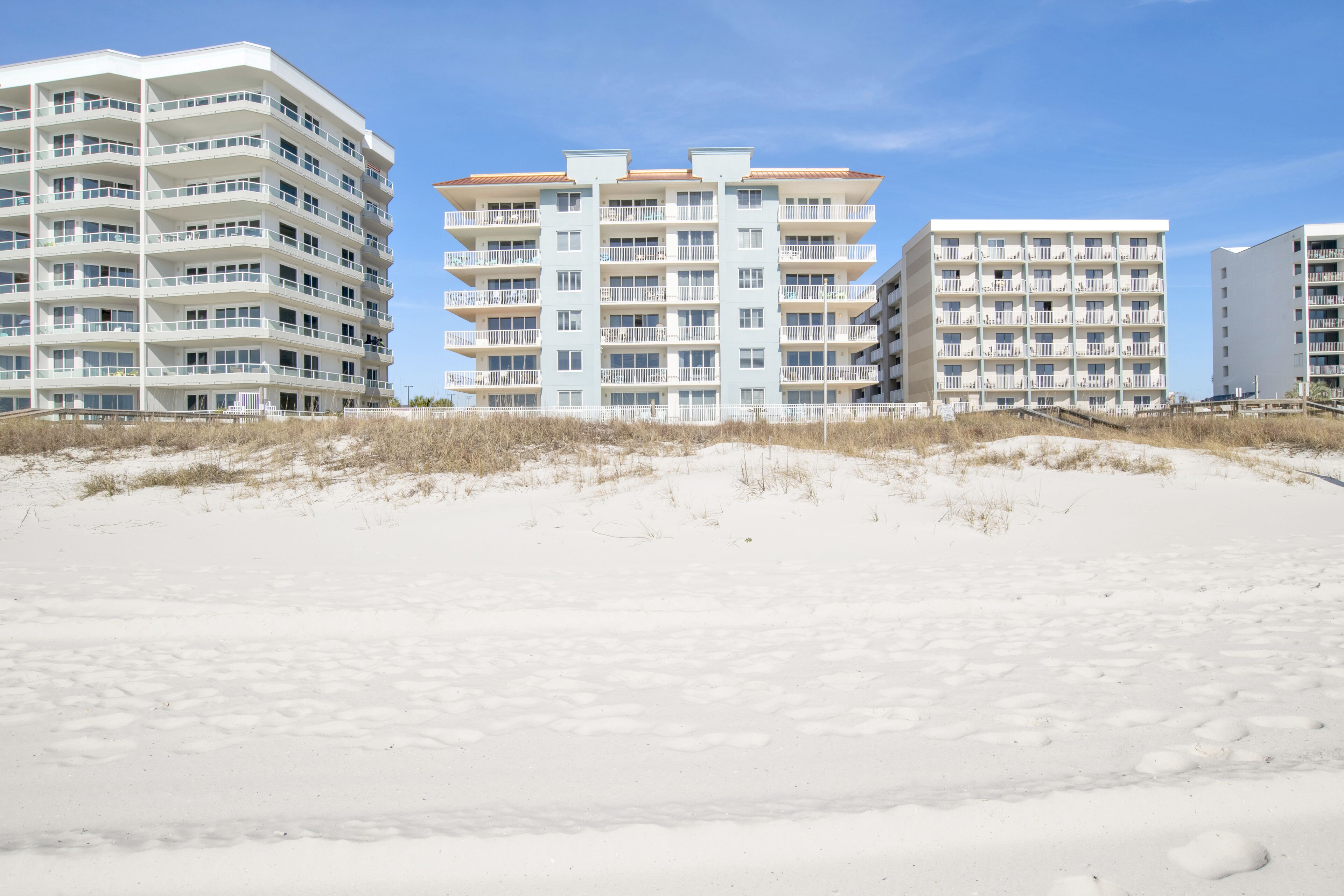Un hotel frente a la playa con varios pisos, balcones y una playa de arena en primer plano.