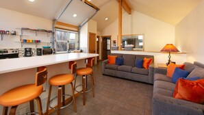 A modern kitchen with a central island, bar stools, a sofa with orange and blue cushions, and a lamp.