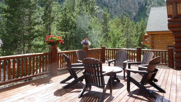 Um deck de madeira com cadeiras Adirondack e uma mesa, com vista para uma montanha arborizada.