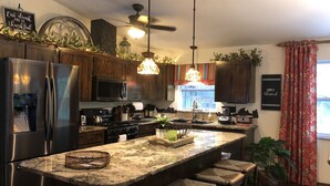 A modern kitchen with a stainless steel refrigerator, a microwave, a stove, and a sink. There are wooden cabinets, a large island with bar stools, and various kitchen appliances. - A SLICE OF "OLD FLORIDA" AMONG GRANDFATHER OAKS. (Crystal River)