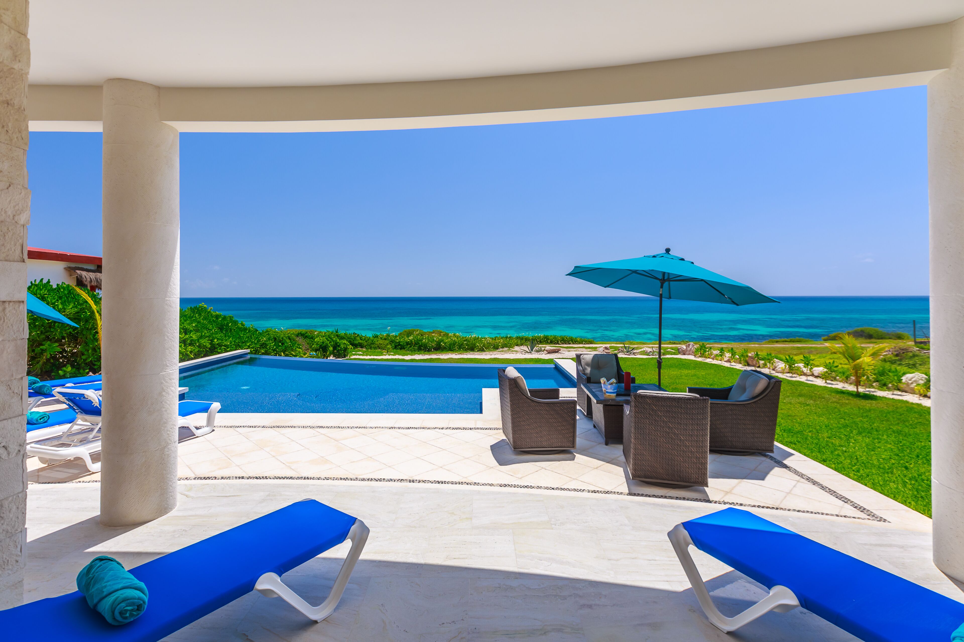 A poolside area with lounge chairs, a table, and an umbrella, overlooking a clear blue ocean.