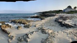 A coastal landscape with rocky terrain, clear water, and a building with a white facade and dark roof. - Casa Flor de Mayo  5BR Villa with Infinity Pool & Stunning Ocean Views (Isla Mujeres)