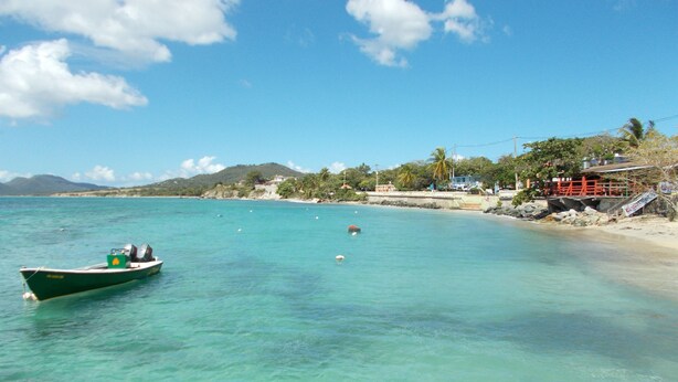 A boat on a clear turquoise sea with a beach and buildings in the background. - Dos Palmas-3Bdrm, 2Ba, Walk One Block to Caribbean,Malecon,restaurants,etc (Vieques)