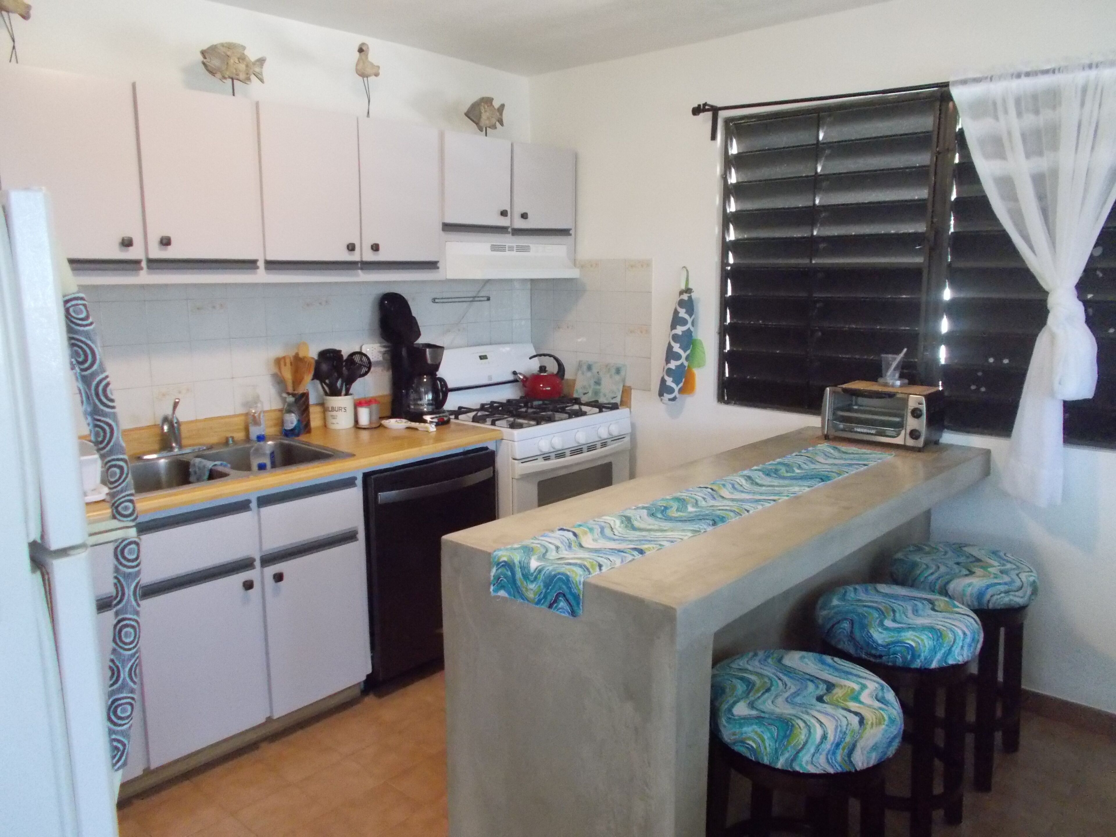 A compact kitchen with white cabinets, a gas stove, a coffee maker, and a toaster. There are stools at the counter and a window with blinds.