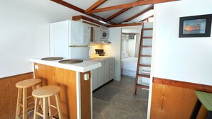 Een compacte keuken met een centraal eiland, houten krukken, en een bed dat in de aangrenzende kamer zichtbaar is.