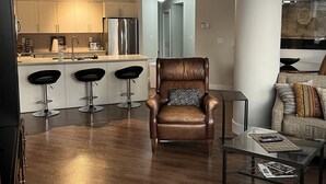 A modern living room with a brown leather armchair, a glass coffee table, and a sofa with patterned cushions. A kitchen with white cabinets and stainless steel appliances is visible in the background. - Beautiful Downtown Executive Suite (Edmonton)