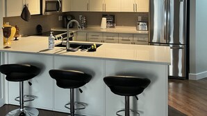 A modern kitchen with white cabinets, a stainless steel refrigerator, a microwave, and a coffee machine. There are three black bar stools in front of the kitchen island. - Beautiful Downtown Executive Suite (Edmonton)