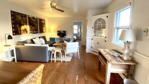 A spacious living room with a grey sofa, a small white table, and a wooden bench. There is a ceiling fan, a TV, and a window with a view of the outside. - Secluded 1 Br Cottage on the Lake, Close to Ditch with OS Shower & Fabulous Yard (Montauk)