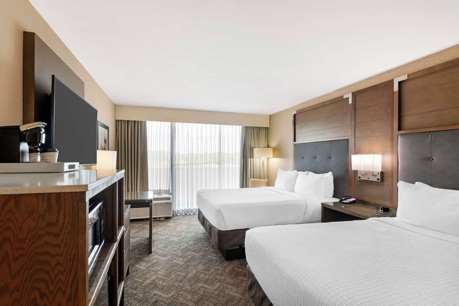 A hotel room with two beds, a desk with a coffee maker, a TV, and a large window with curtains.