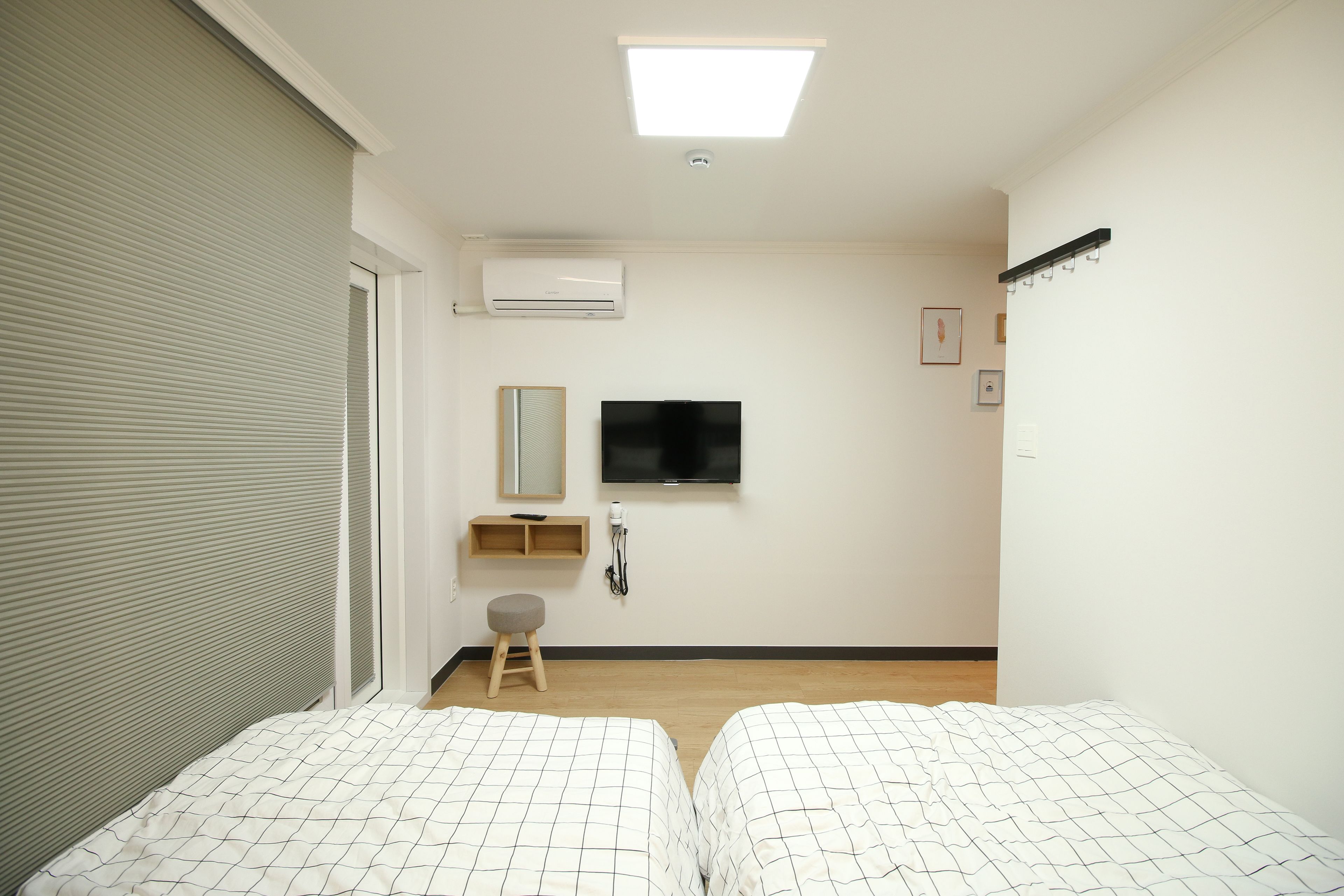 A hotel room with two beds, a flat-screen TV, a small stool, a mirror, and a shelf with a hairdryer.
