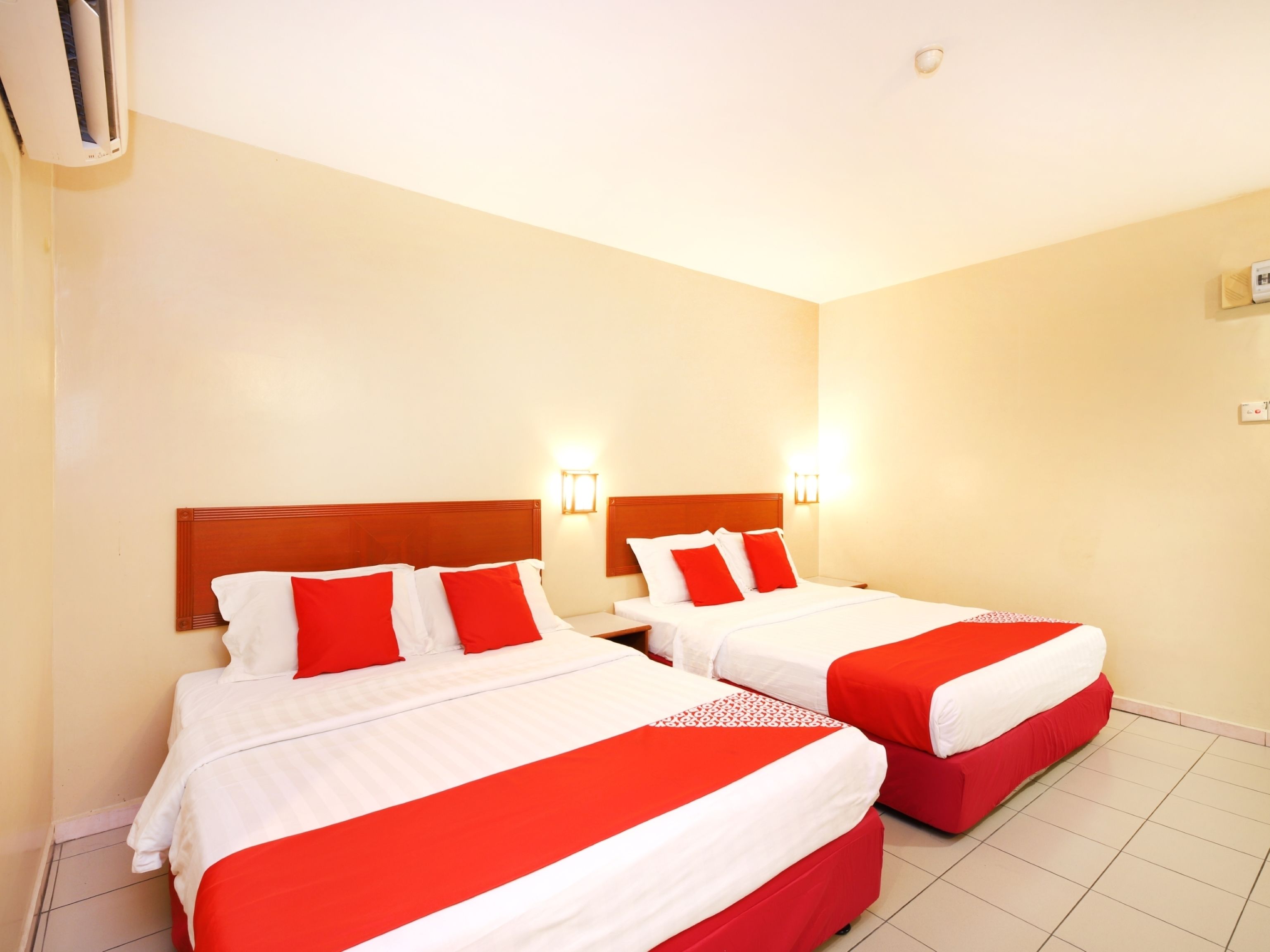 Une chambre d’hôtel avec deux lits, chacun doté d’une literie rouge et blanche, et d’une tête de lit en bois. Des appliques murales sont fixées au-dessus de chaque lit.