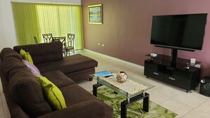 Una sala de estar con un sofá marrón, una mesa de centro de vidrio, un televisor sobre un soporte negro y una alfombra verde estampada.