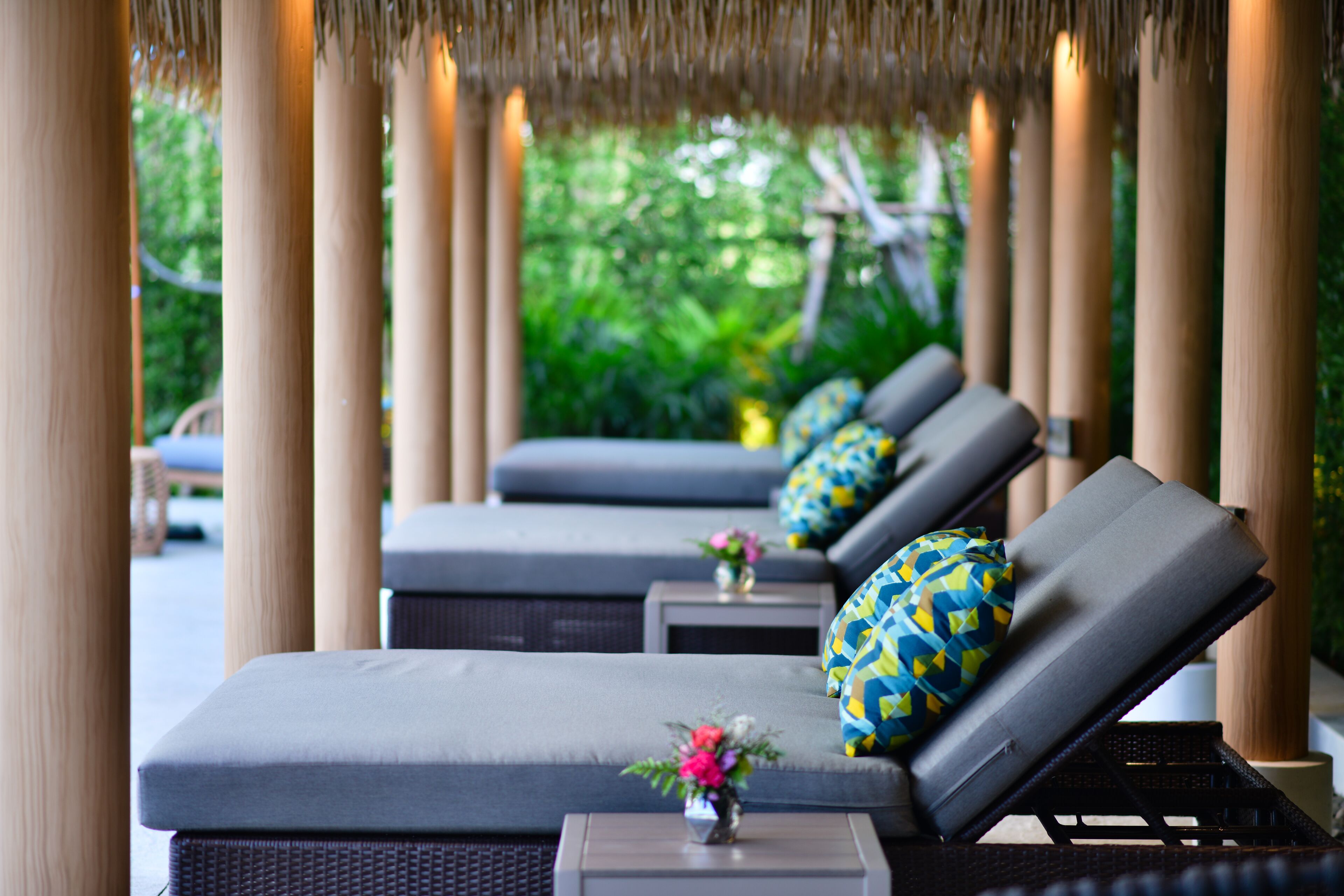 A row of lounge chairs with cushions, a small table with a vase of flowers, and a thatched roof structure.
