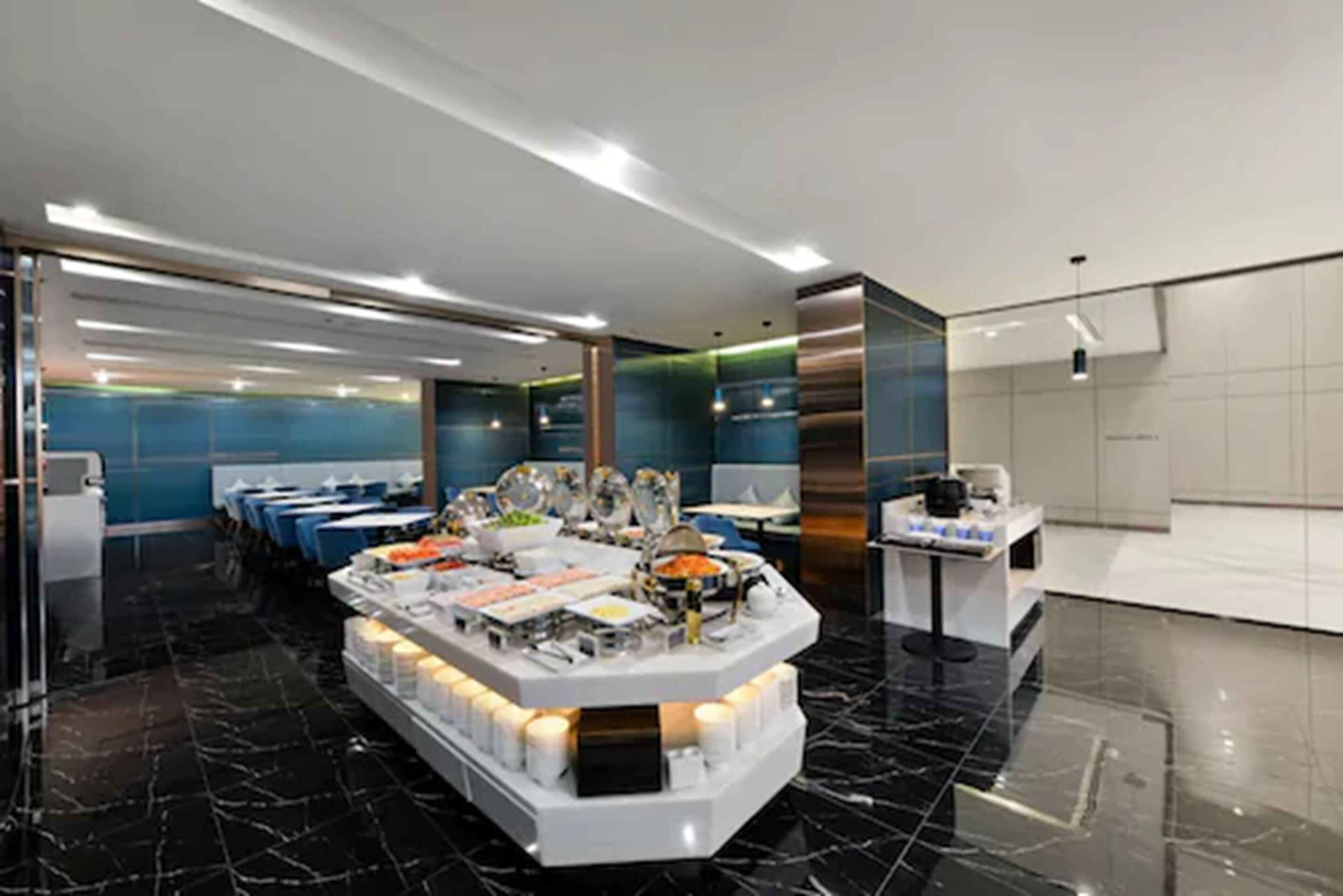 A modern hotel lobby with a central buffet area, seating arrangements, and a reception desk.