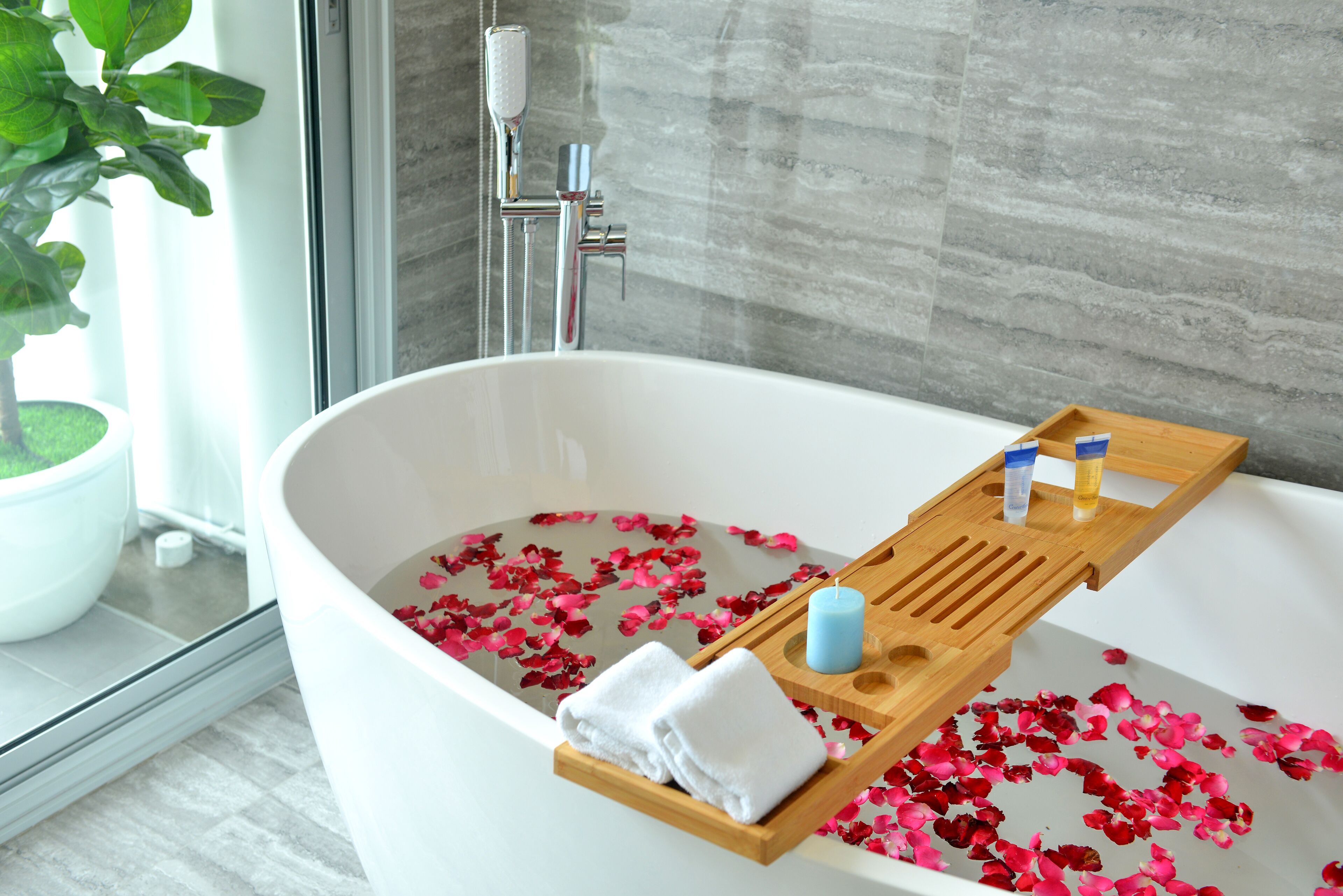 A modern bathroom with a freestanding bathtub filled with rose petals, a wooden tray with toiletries, and a potted plant by the window.