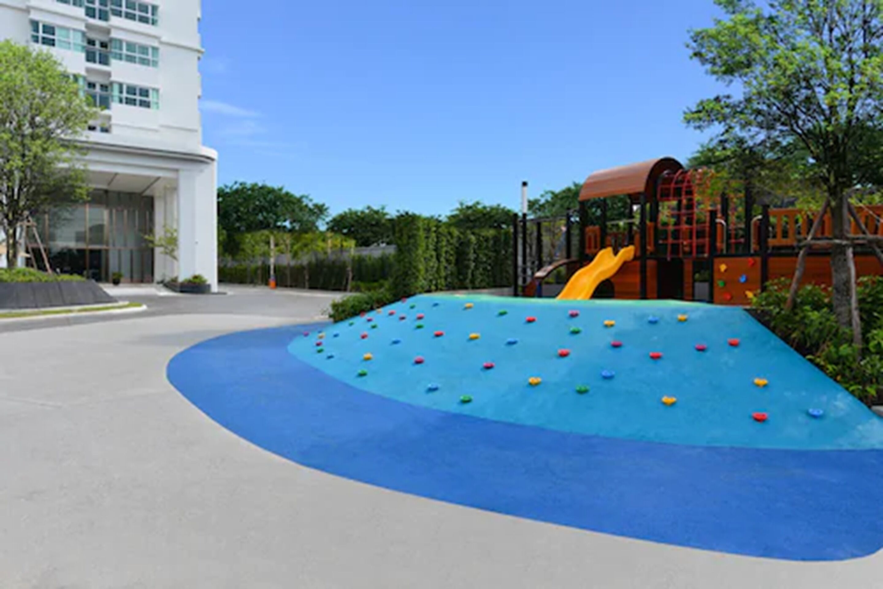 A playground with climbing structures and a slide, surrounded by a building and greenery.