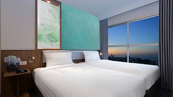 A hotel room with a large bed, a nightstand with a vase of flowers, and a view of the city at sunset.