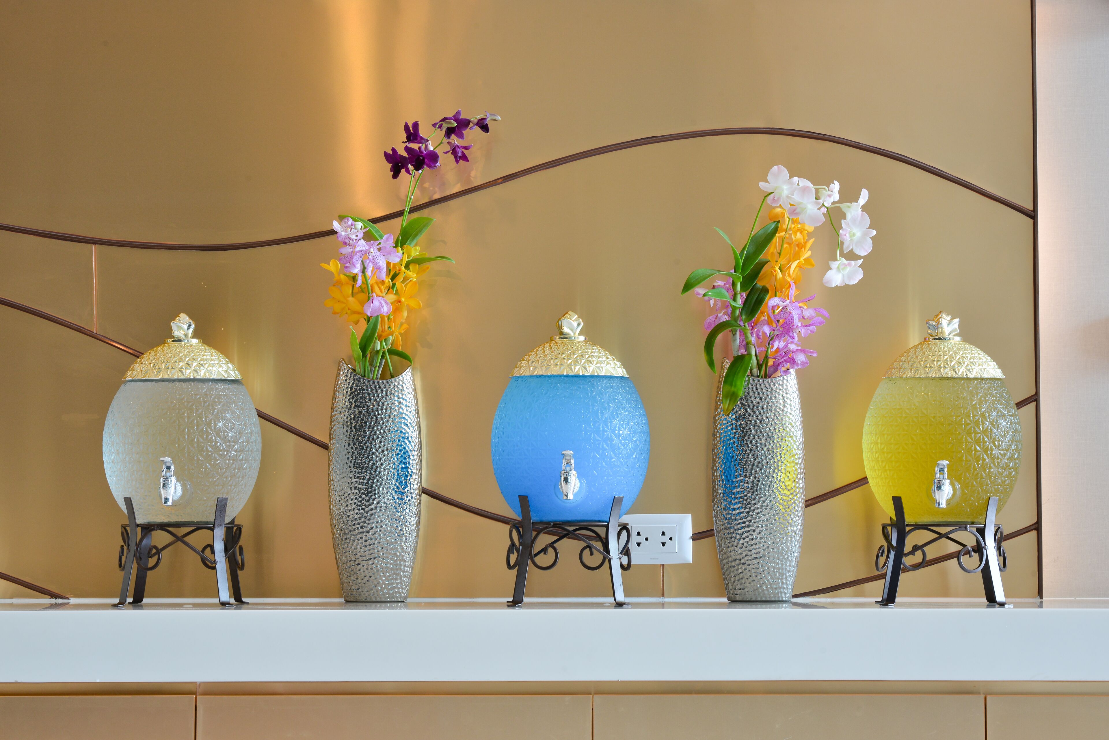 Five glass vases with flowers on a white shelf against a wall.
