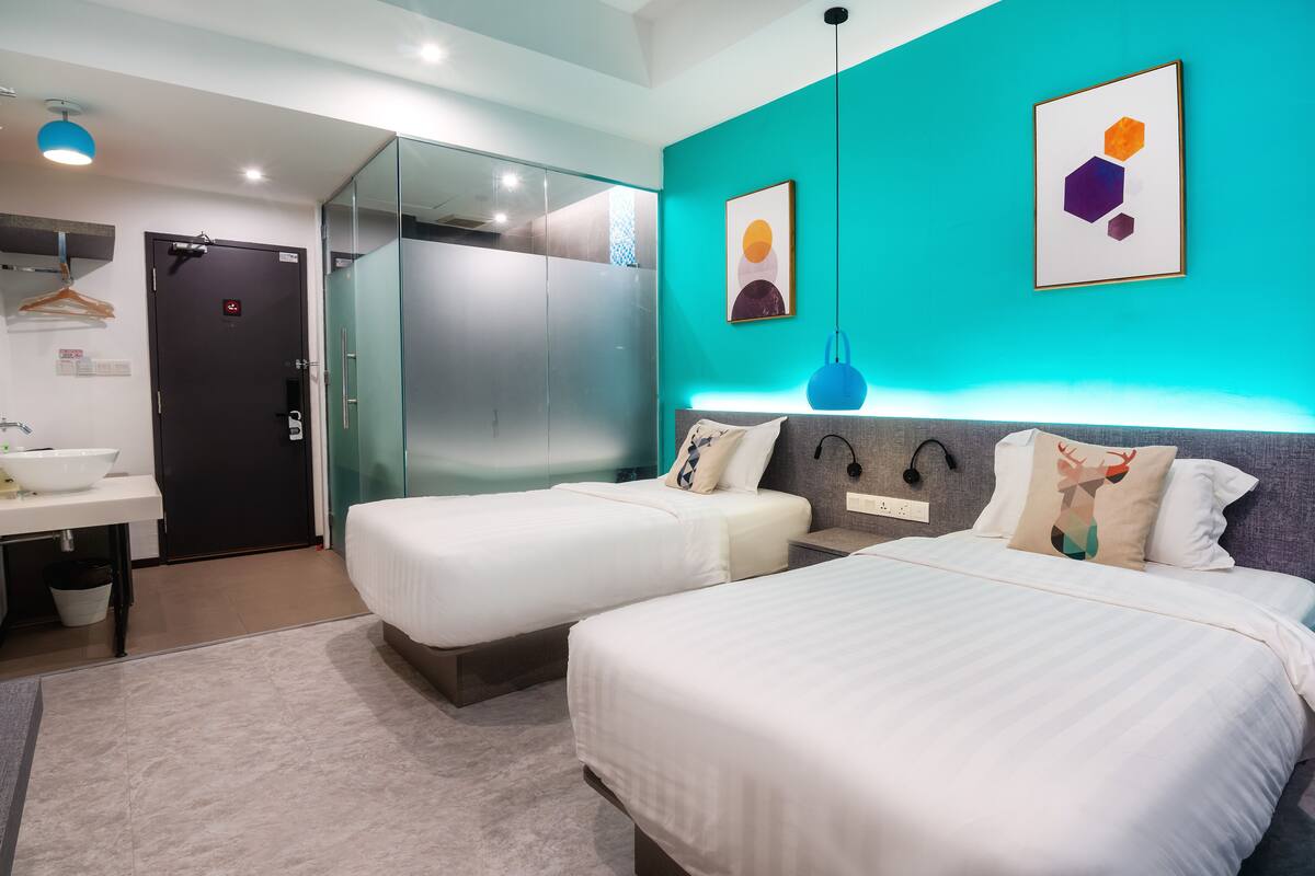 Quarto de hotel moderno com duas camas de solteiro, banheiro com pia e espelho, guarda-roupa e uma parede verde-água com arte abstrata.