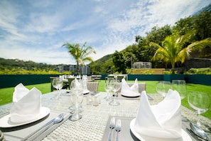 An outdoor dining area with tables set for a meal, surrounded by lush greenery and a view of a hill. - Oceana Resort Phuket (Kamala)