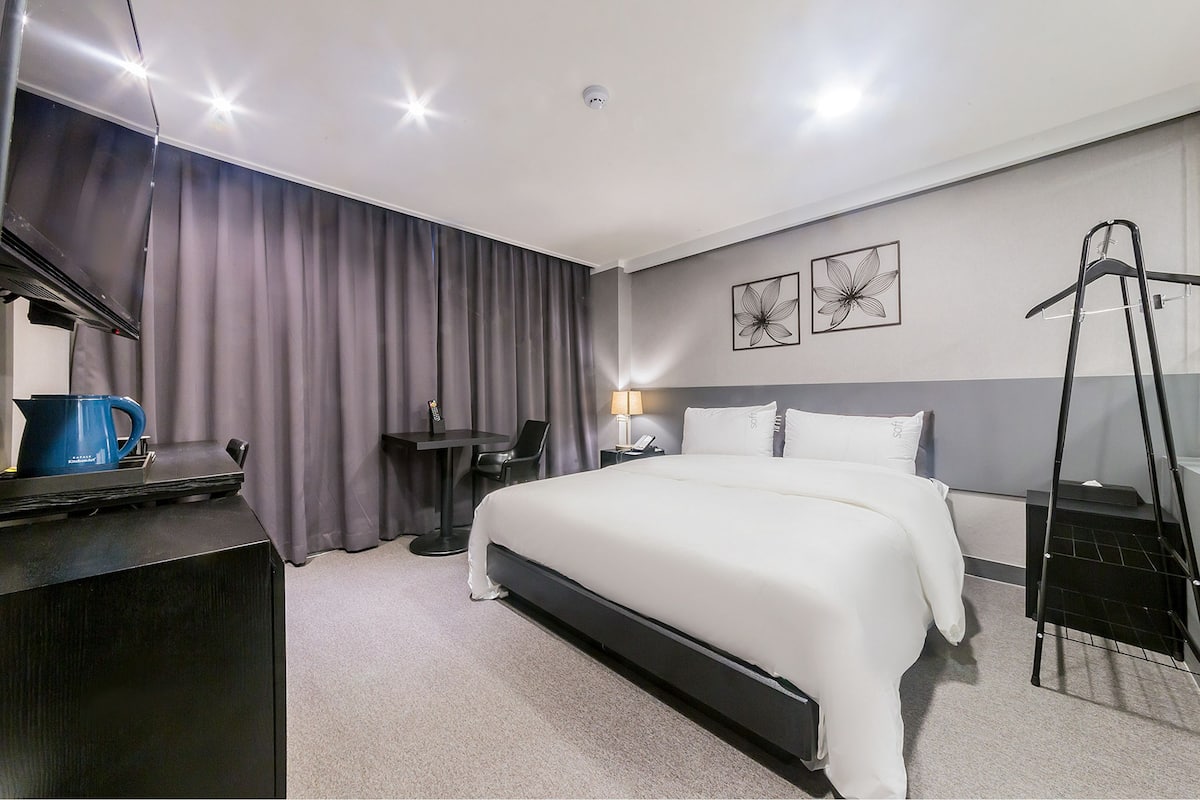 A hotel room with a bed, a desk, a chair, a coffee machine, a lamp, and a coat rack.