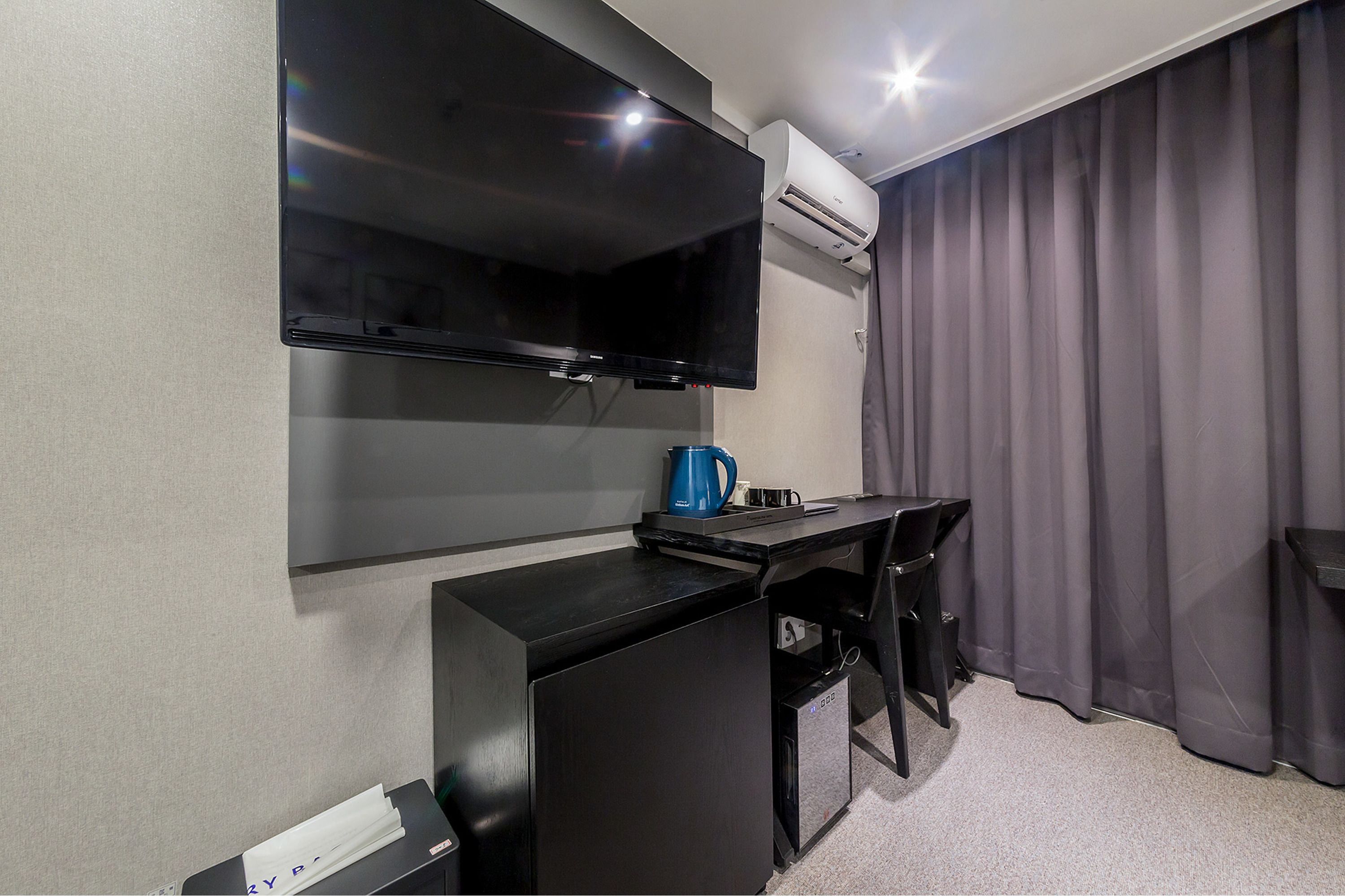Ein kompaktes Hotelzimmer mit Flachbildfernseher an der Wand, einem kleinen Schreibtisch mit Stuhl, einer Kaffeemaschine und einem Mülleimer.