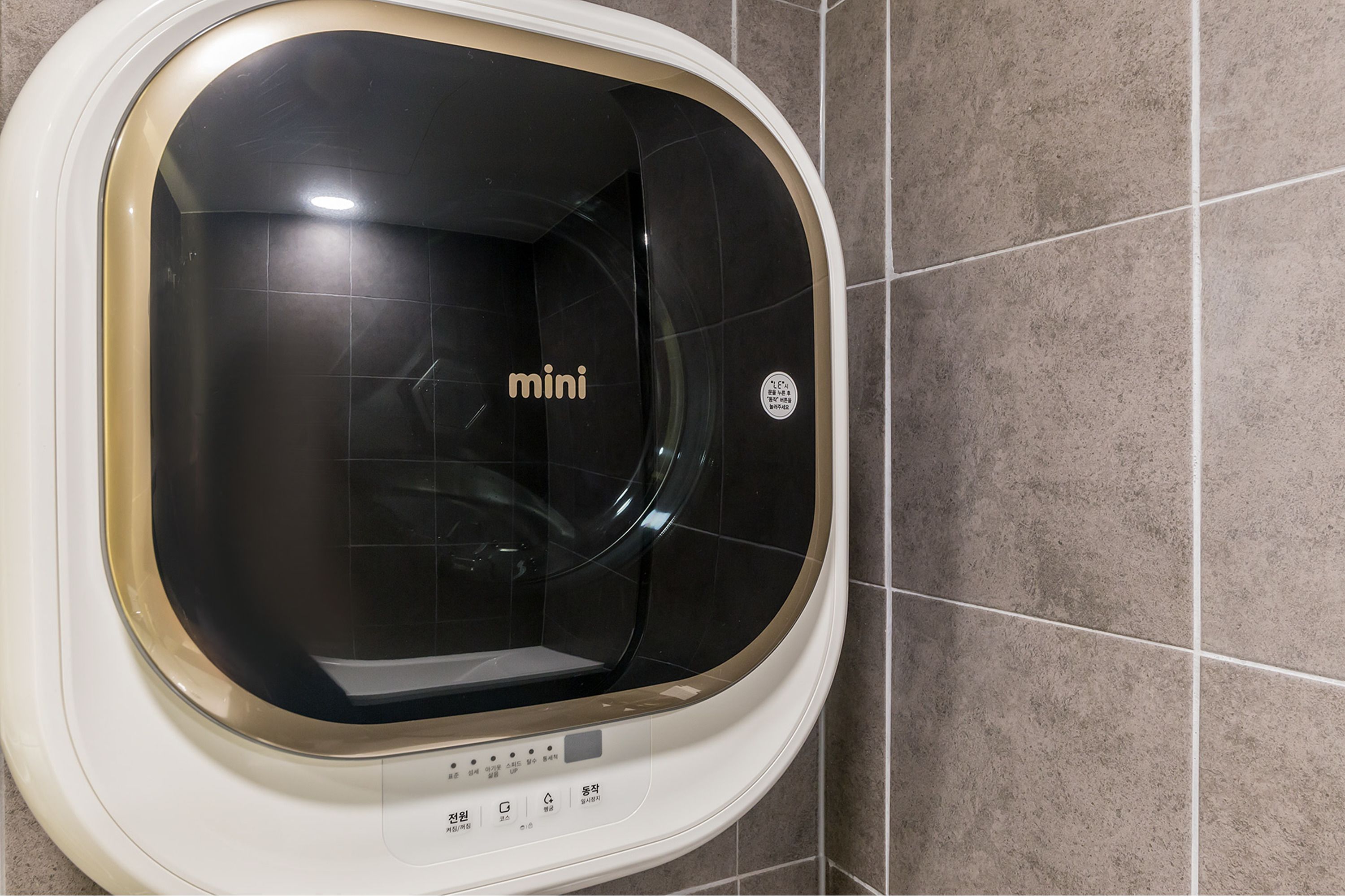 A compact, modern washing machine with a control panel, set against a tiled wall.
