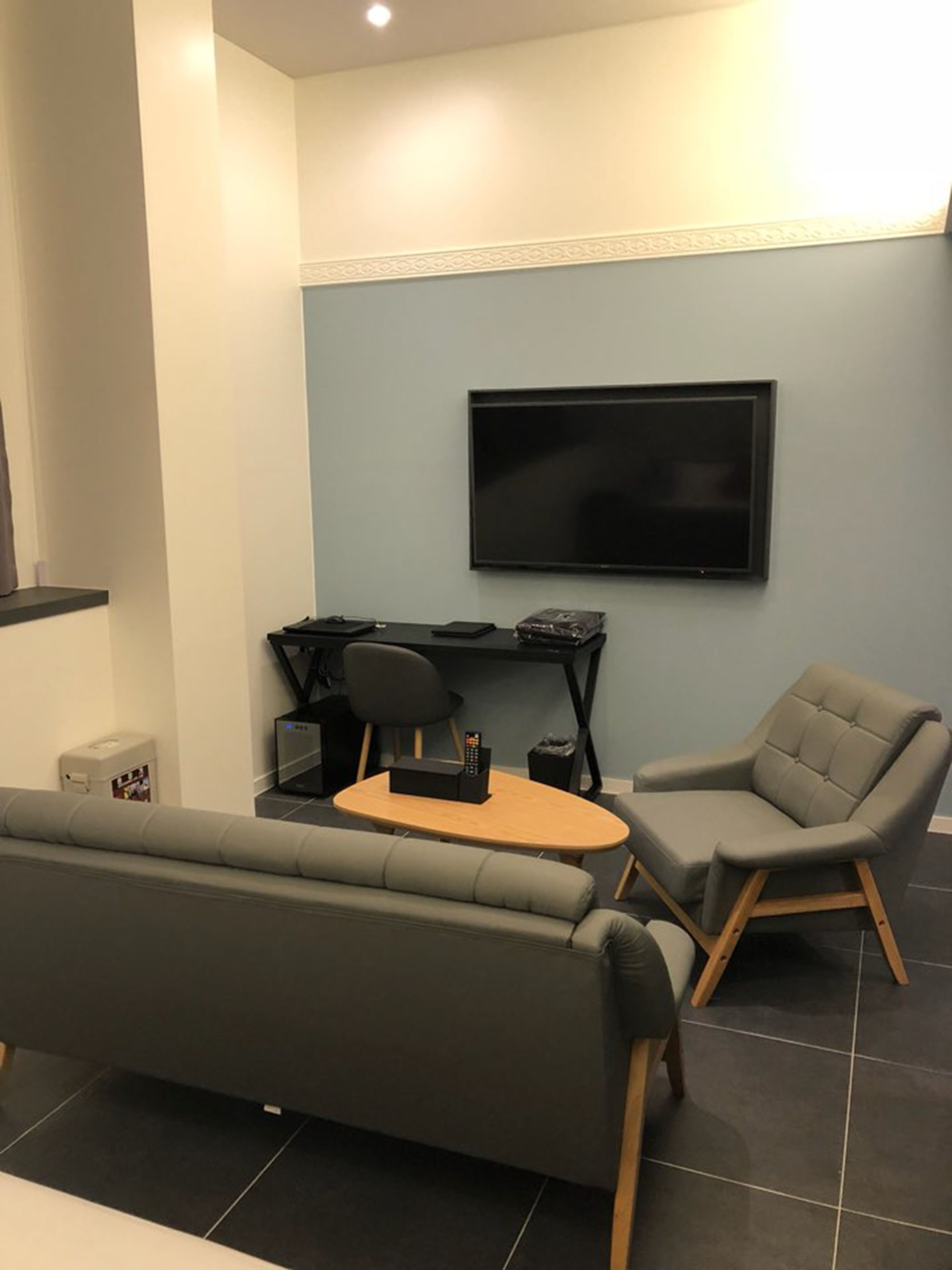 A modern living room with a grey sofa, a small wooden table, a grey armchair, and a flat-screen TV mounted on the wall.