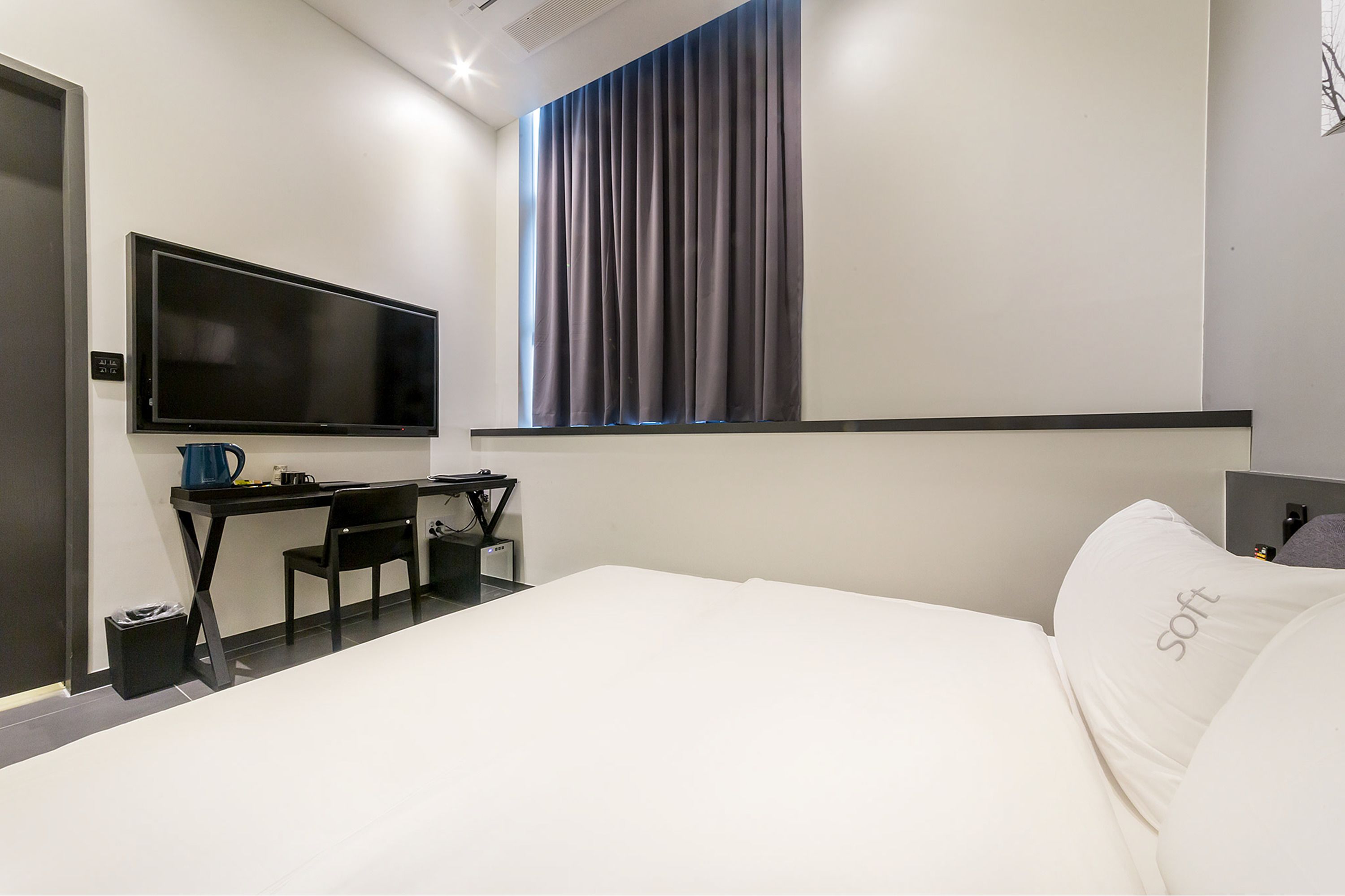 A modern hotel room with a large bed, a flat-screen TV mounted on the wall, a desk with a chair, and a window with curtains.