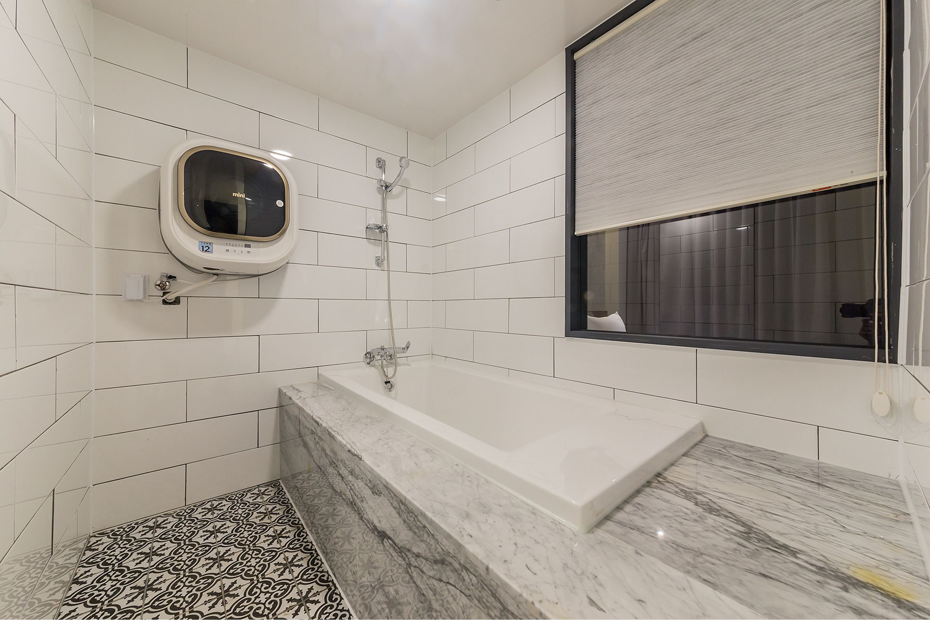 Une salle de bain moderne avec des carreaux blancs, une baignoire en marbre, une douchette à main et une fenêtre avec des stores.