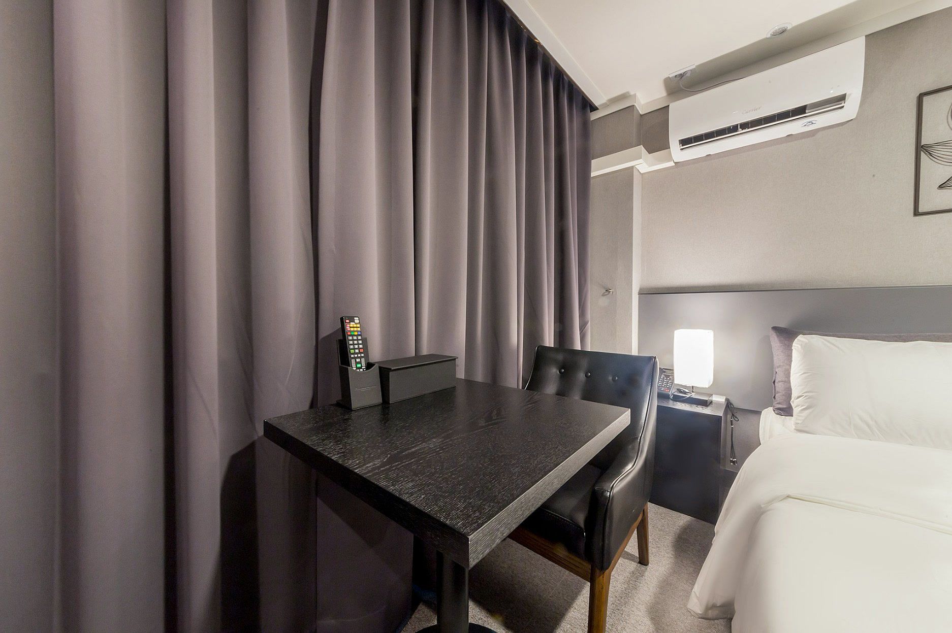 A hotel room with a bed, a desk with a chair, a bedside table with a lamp, and a window with curtains.