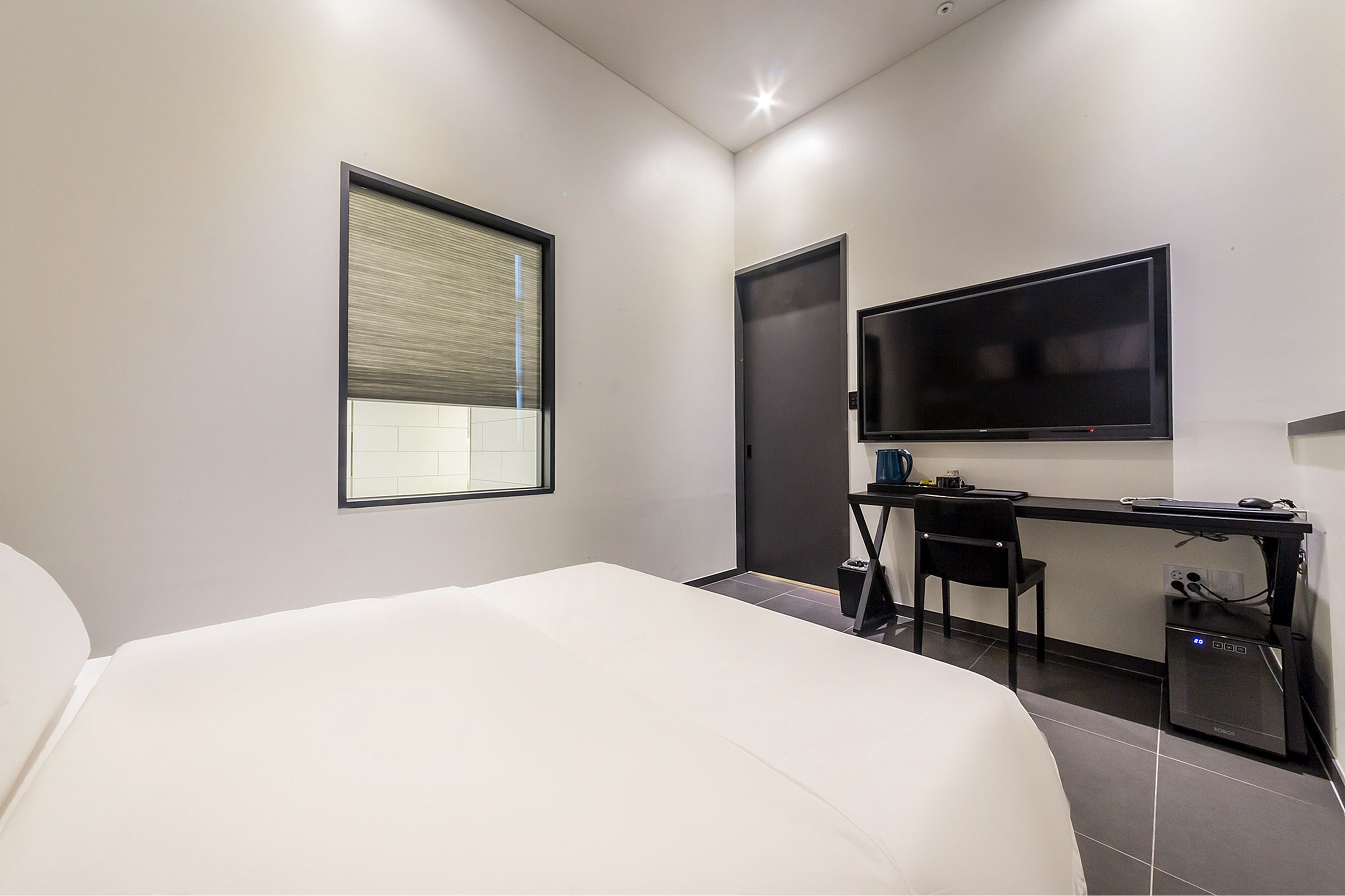 A modern hotel room with a large bed, a desk with a computer, a flat-screen TV, and a window with blinds.
