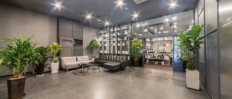 A modern lobby with a grey sofa, a coffee table, and potted plants. There is a glass display case with items and a door leading to a kitchen area.
