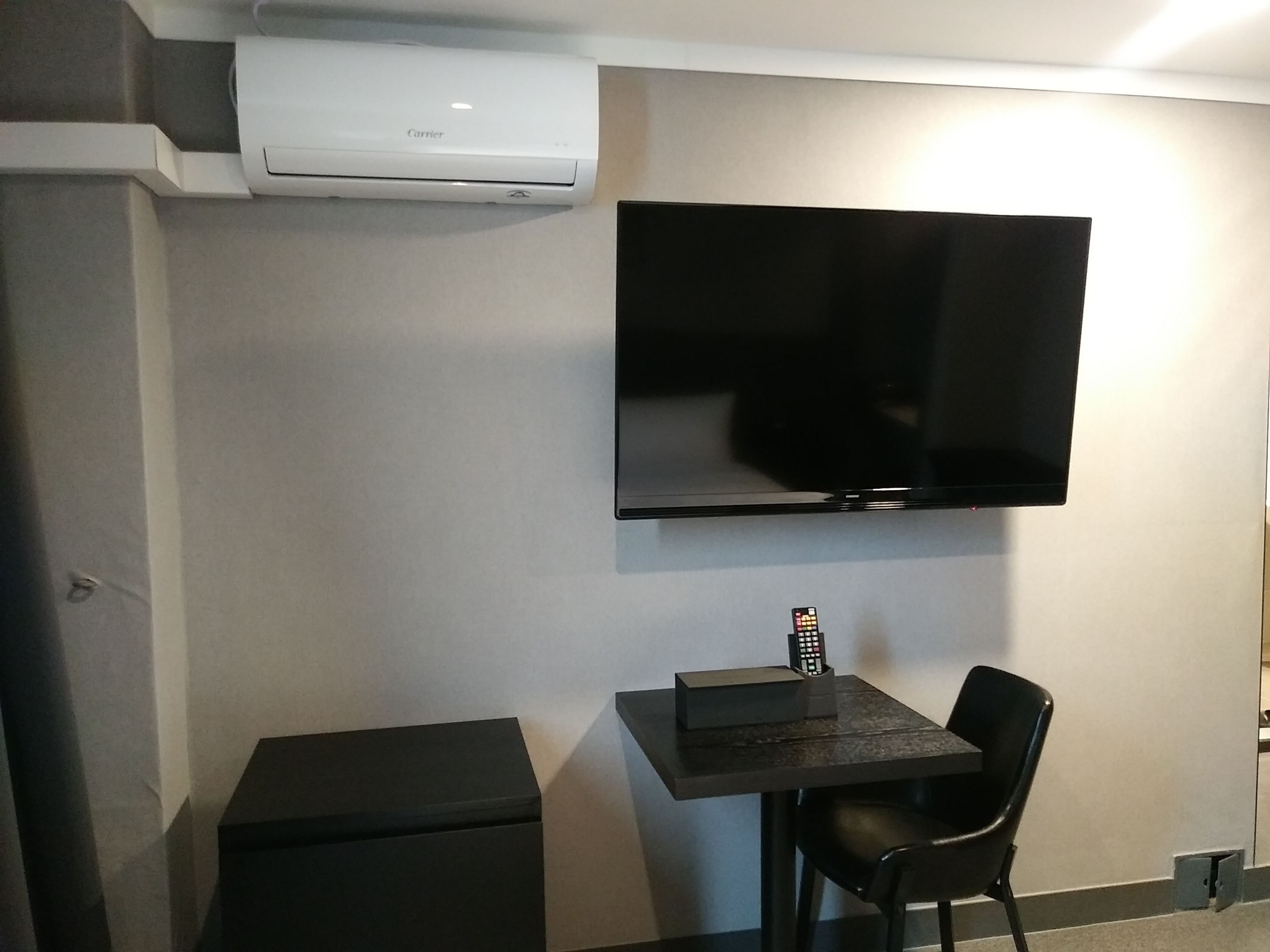 Una habitación con una televisión de pantalla plana montada, una pequeña mesa con un control remoto, una silla negra y un mueble de almacenamiento negro compacto.