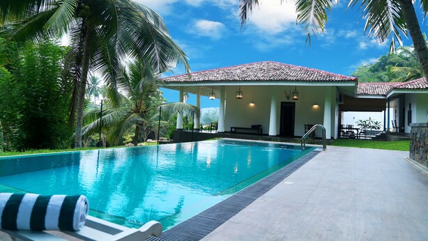 A swimming pool with a clear blue water, surrounded by palm trees and a building with a tiled roof. - Thompson Manor - A Luxury Villa in Galle (Akmeemana)