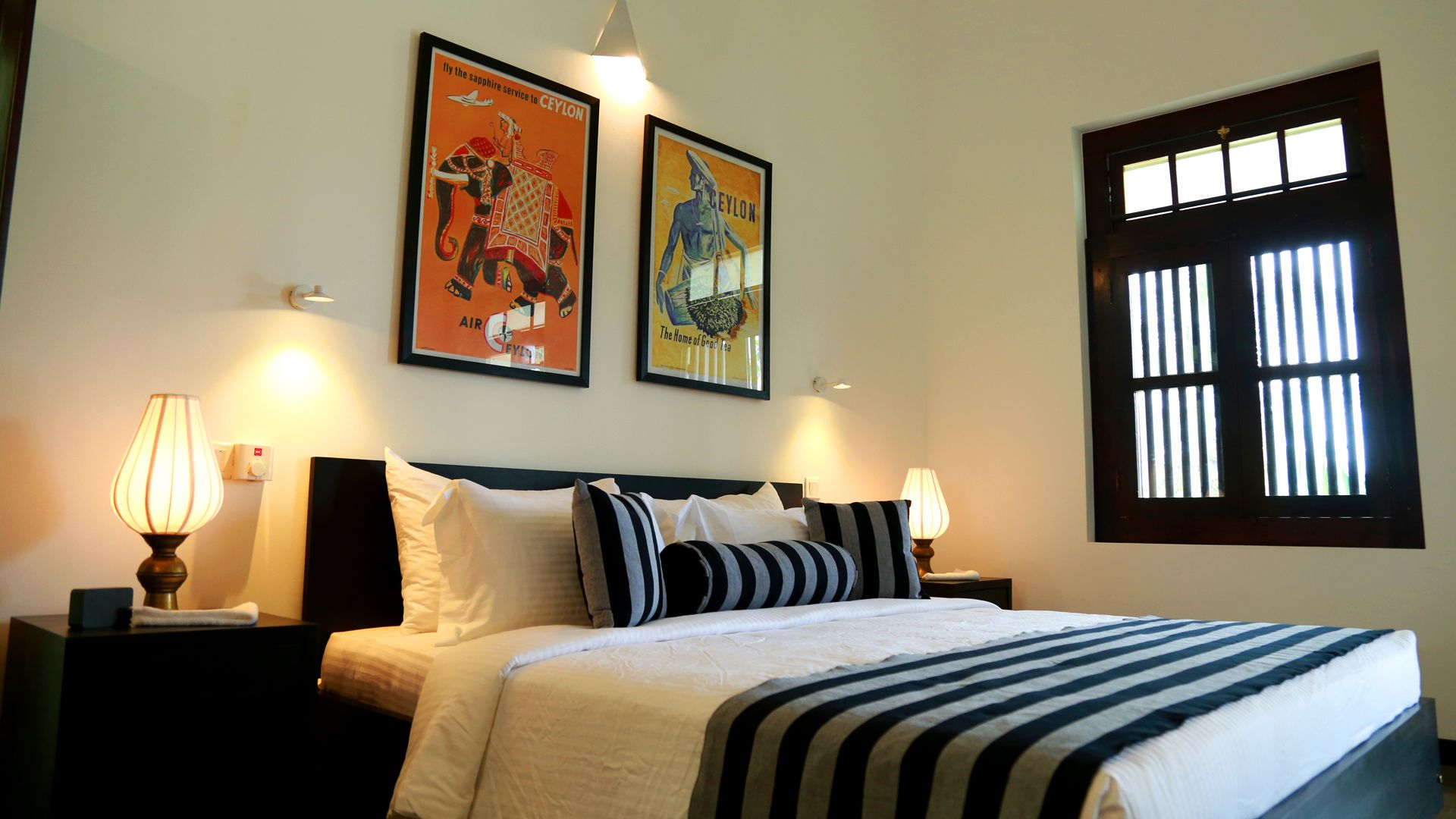 A hotel room with a bed, two wall-mounted lamps, a window with bars, and two framed posters on the wall.