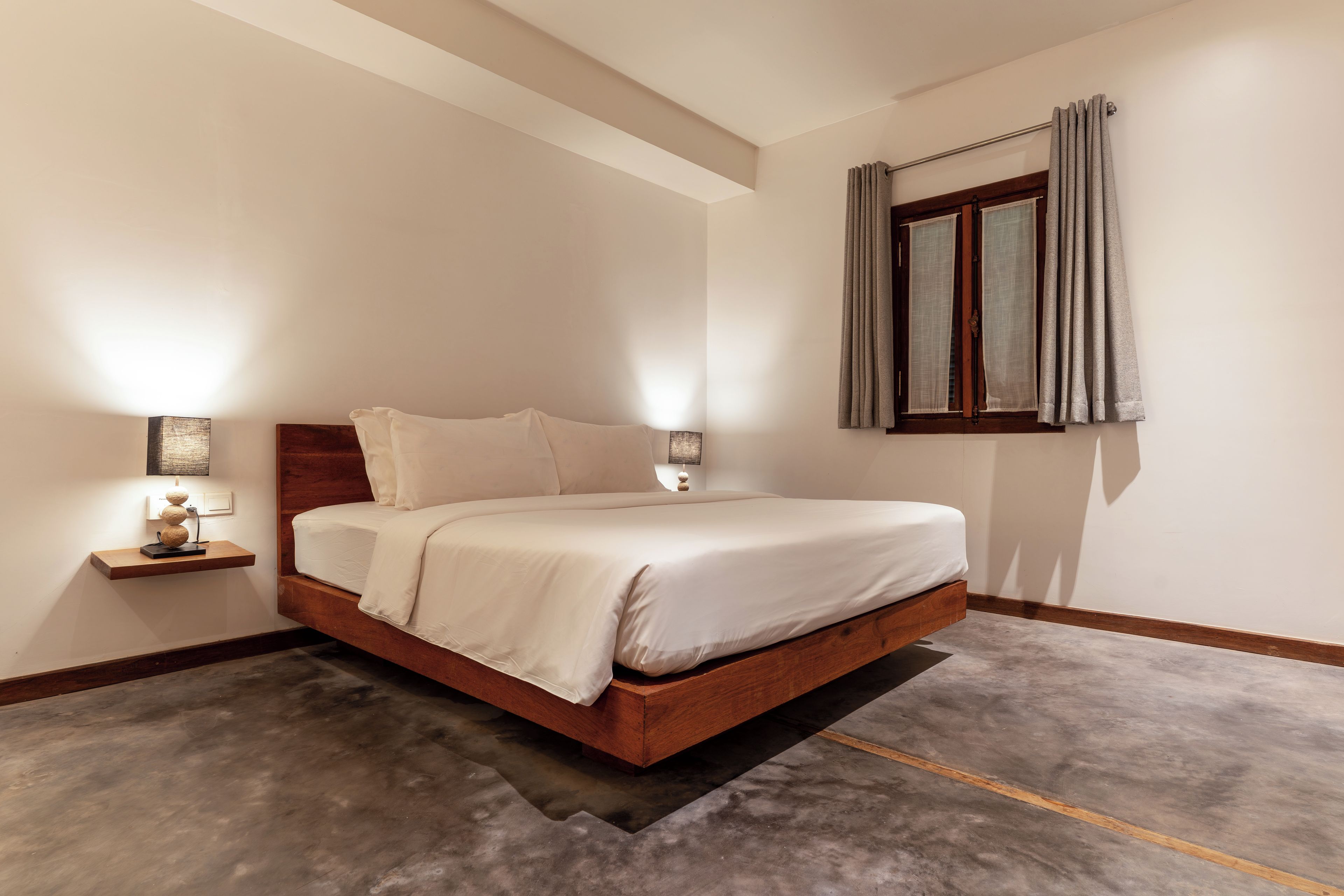 A bedroom with a wooden bed frame, a nightstand, a wall-mounted lamp, and a window with curtains.