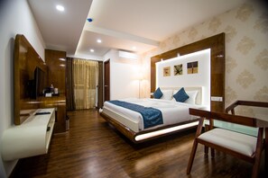 A hotel room with a wooden floor, a bed with a white and blue bedspread, a wooden headboard, a nightstand, a desk with a chair, a mirror, and a window with curtains. - The White Tree Executive Suites (Bengaluru)