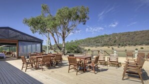 Een houten terras met buitenmeubilair, een glazen uitbouw en uitzicht op een rivier en heuvels.