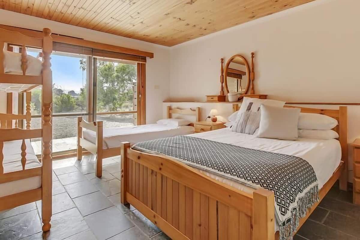 A bedroom with a wooden bed, a nightstand, a mirror, and a window with a view of greenery.