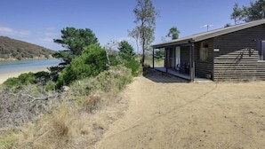 Een rustieke hut met een veranda, gelegen op een zandig gebied met uitzicht op een strand en heuvels op de achtergrond.