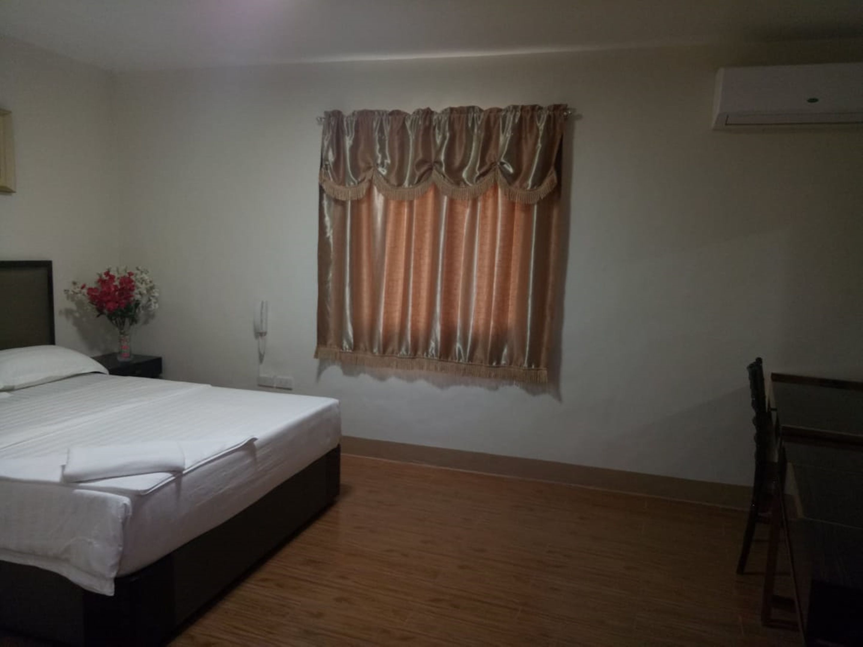 Un dormitorio con cama, mesita de noche con un jarrón de flores, ventana con cortinas, una silla y un equipo de aire acondicionado.