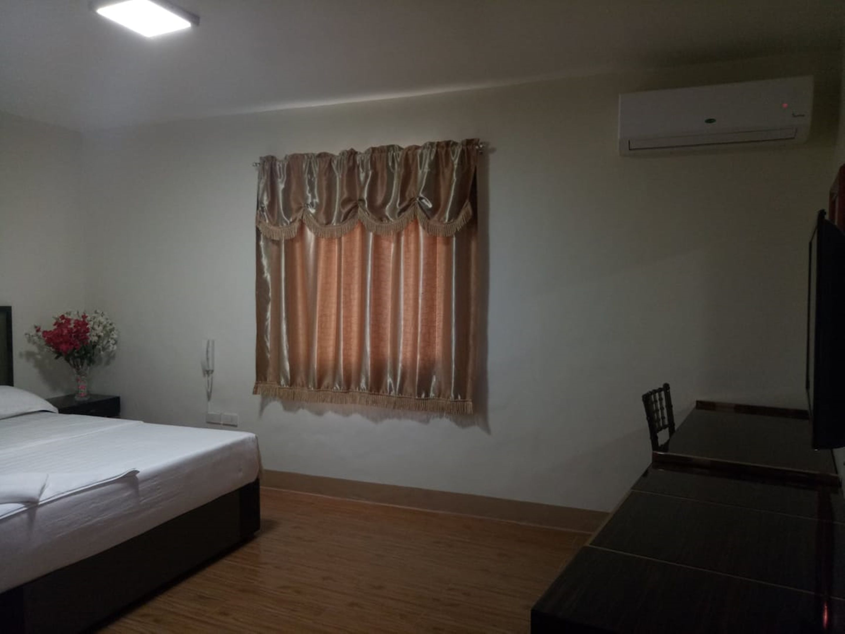 A hotel room with a bed, a small table, a chair, a nightstand, a vase with flowers, and an air conditioning unit.