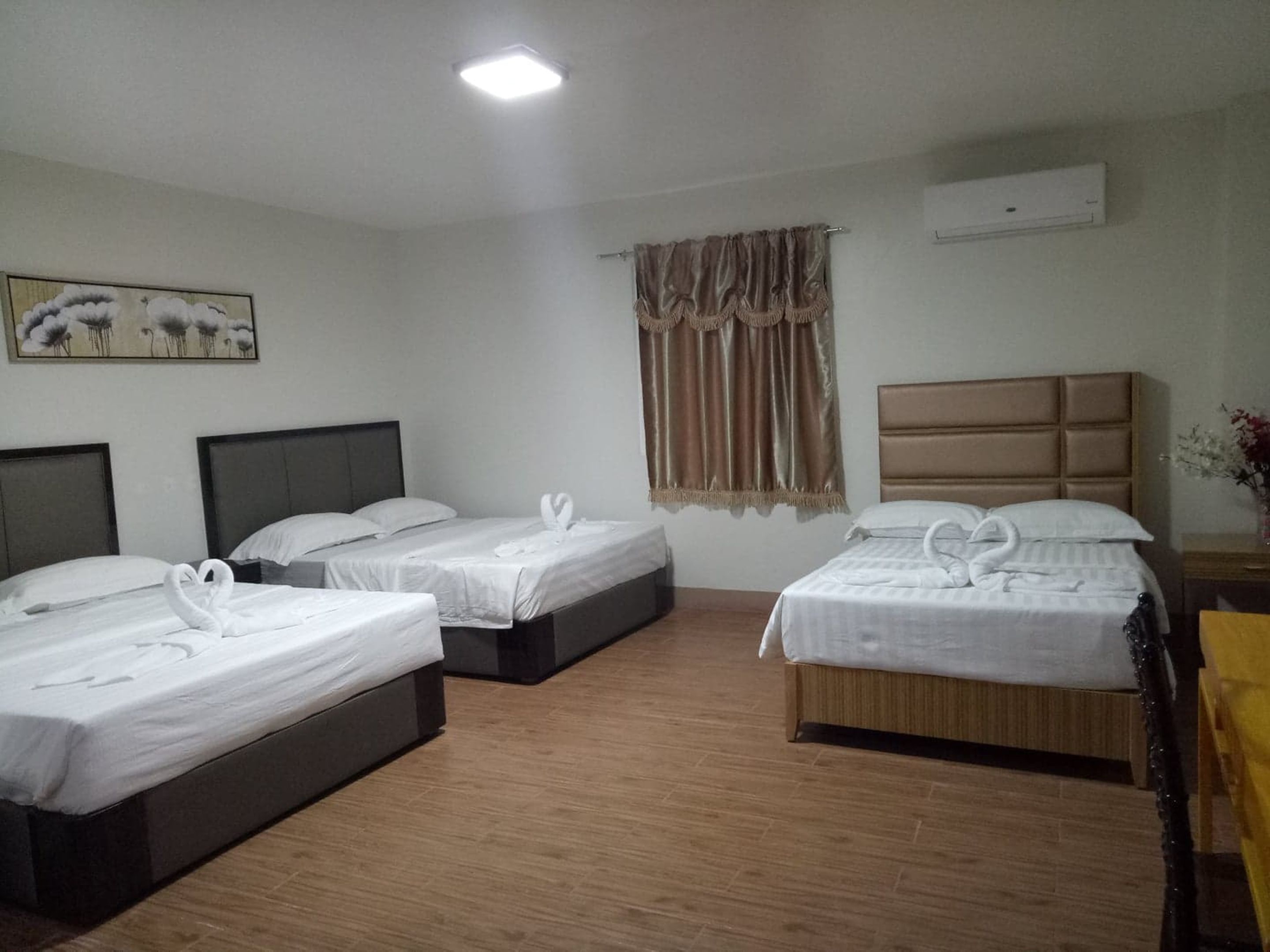 Habitación de hotel con tres camas, cada una con ropa de cama blanca y toallas con forma de cisne decorativas. Hay un cuadro en la pared, una ventana con cortinas y un equipo de aire acondicionado también en la pared.